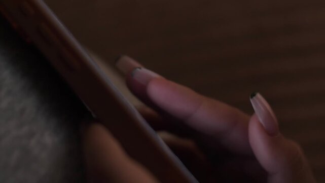 A woman's finger with long French manicure quickly scrolls through a smartphone screen. Close-up shot of interacting with a touchscreen device in a dark room.