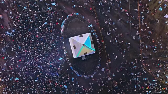 An extended aerial drone orbit circles the illuminated top of the Obelisco in Buenos Aires on July 19, 2026, as clouds of smoke drift past and sparks scatter across the crowd below.