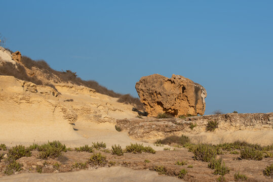 A single sandstone rock lies on the slope of a mountain. In the background is a blue sky. In the foreground is sparse vegetation.