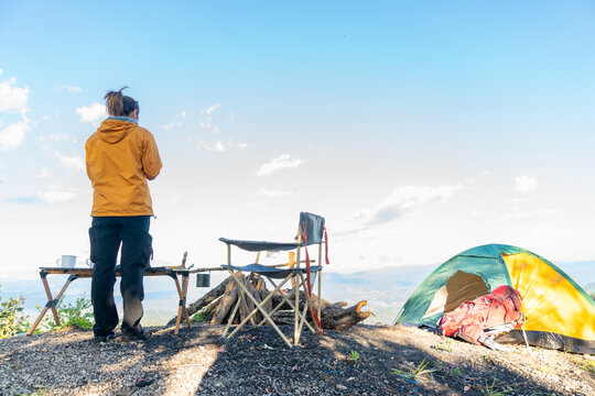 Asian woman drinking coffee while solo travel nature and camping at forest mountain. Female backpacker relaxing at campsite, social detox, enjoy nature therapy morning beverage with outdoor lifestyle