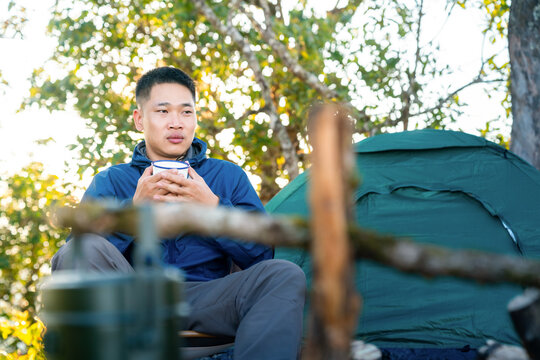 Asian man drinking hot coffee while solo travel nature and camping at forest mountain. Male backpacker relaxing at campsite, social detox, enjoy nature therapy morning beverage with outdoor lifestyle