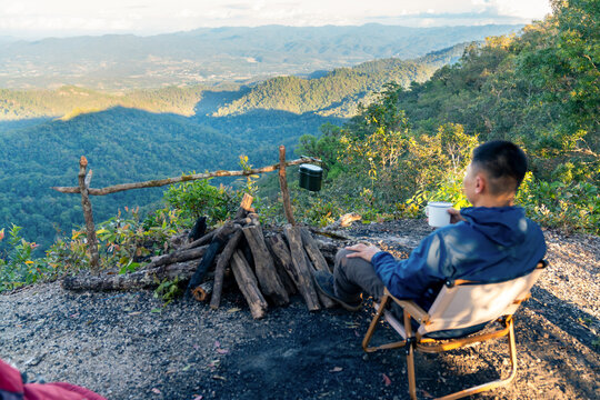 Asian man drinking hot coffee while solo travel nature and camping at forest mountain. Male backpacker relaxing at campsite, social detox, enjoy nature therapy morning beverage with outdoor lifestyle