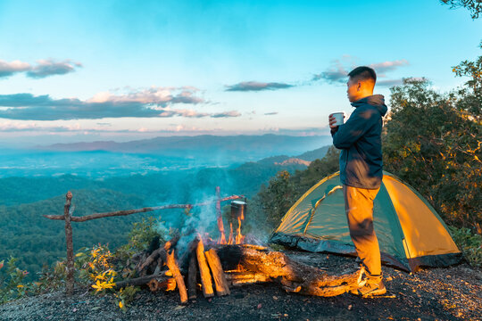 Asian man drinking hot coffee while solo travel nature and camping at forest mountain. Male backpacker relaxing at campsite, social detox, enjoy nature therapy morning beverage with outdoor lifestyle