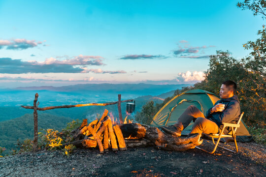 Asian man drinking hot coffee while solo travel nature and camping at forest mountain. Male backpacker relaxing at campsite, social detox, enjoy nature therapy morning beverage with outdoor lifestyle