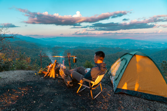 Asian man drinking hot coffee while solo travel nature and camping at forest mountain. Male backpacker relaxing at campsite, social detox, enjoy nature therapy morning beverage with outdoor lifestyle