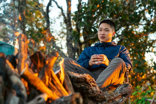 Asian man drinking hot coffee while solo travel nature and camping at forest mountain. Male backpacker relaxing at campsite, social detox, enjoy nature therapy morning beverage with outdoor lifestyle