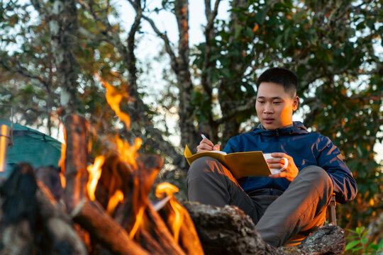 Asian man writing travel journal and drinking hot coffee beside campfire and tent in forest mountain. People relax and enjoy travel outdoor active lifestyle hiking, backpacking and nature healing.