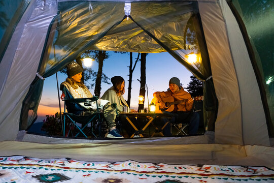 Group of Adult friends camping on mountain, drinking beer and playing guitar together by tent. Man and woman enjoy outdoor active lifestyle campsite leisure and friendship travel on holiday vacation.