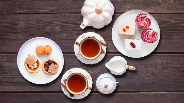 Fall theme afternoon tea table stop motion animation placement on a dark wood background. Pumpkin and apple theme savory finger foods, sandwiches, scones, and desserts. Above view.