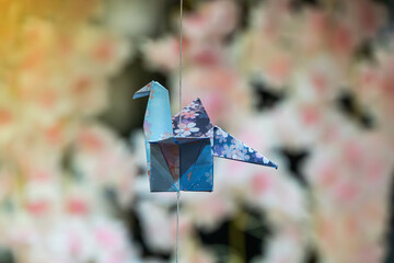 Naklejka premium Origami crane suspended by wire against floral background, intricate folded paper bird, light blue and dark blue with subtle floral pattern, soft pastel colors, gentle diffused lighting, elegant
