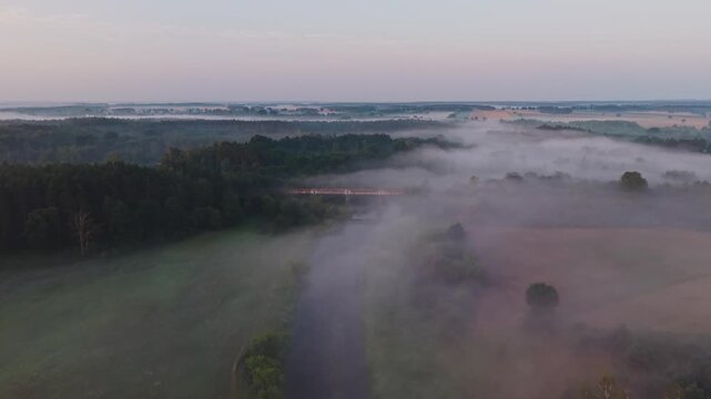 Aerial view of a rusted metal bridge crossing the misty Wkra river surrounded by forest and fields at dawn