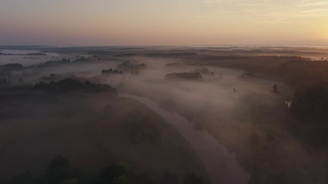 Aerial view of a narrow river winding through fields and forest under a thick layer of morning fog at sunrise
