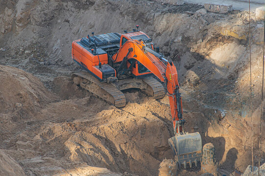 Excavator engaged in earth-moving operations within a large excavation pit, featuring loose soil and debris. Bright orange excavator with extended arm and bucket covered in dirt, highlighting dynamic