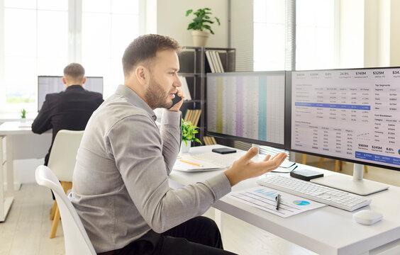 Businessman on phone in office analyzing spreadsheet. He reviews data on computer monitors during a finance call. Manager gestures, overloaded with deadlines. Clear concept of finance analysis.