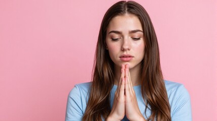 Fototapeta premium Hopeful young woman praying with hands clasped together against a pink background