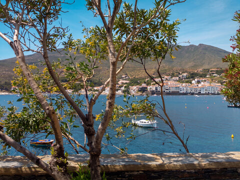 Vue pittoresque d'un village c&ocirc;tier m&eacute;diterran&eacute;en &agrave; travers les branches d'un arbre, avec montagnes en arri&egrave;re-plan et petit port de bateaux tranquille