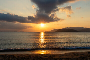 Fototapeta premium Sunset or sunrise over calm ocean waters with silhouetted mountains and dramatic clouds reflecting warm golden light in the sky