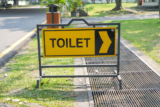 Yellow toilet directional sign with a black arrow mounted on a portable metal stand in a public outdoor area.