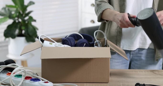 Recycling. Woman putting different broken household appliances into cardboard box at wooden table indoors, closeup