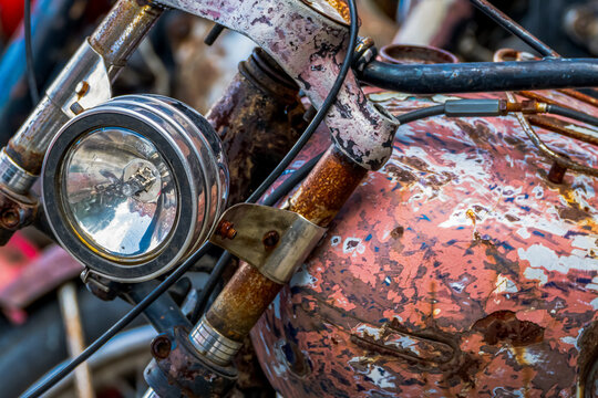 A detailed close-up of a rusty vintage motorcycle, with a round glass headlight and a fuel tank with peeling paint, at a junkyard.