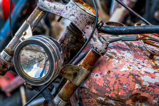 A detailed close-up of a rusty vintage motorcycle, with a round glass headlight and a fuel tank with peeling paint, at a junkyard.