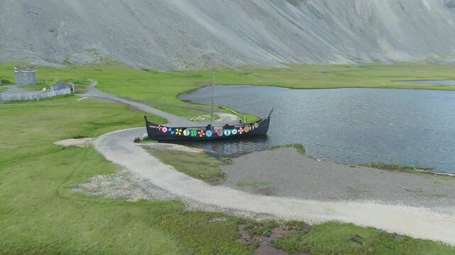 An aerial drone tracking shot pans right along a longship with shields, resting by a stream with a old Viking village film set on Iceland, nestled in a green valley.