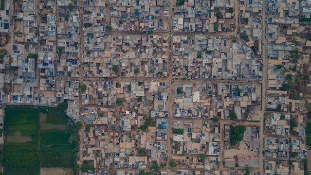 Aerial view of a dense residential neighborhood with flat rooftops, narrow streets, and adjacent agricultural fields in Punjab, Pakistan.