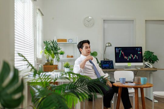 Asian businessman reflecting on stock market trends and investment strategy while working remotely in a bright, plant filled home office with computer monitors displaying trading graphs