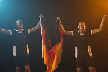 Fototapeta premium African female team wearing black-white jerseys holding black-red-gold flag raising arms onstage