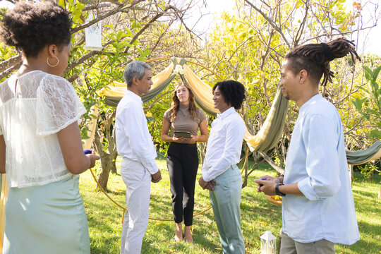 Diverse group with couple and officiant exchanging vows in garden under fabric arch holding tablet