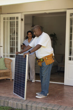 African American pair standing on porch, man in white tee inspecting solar panel with tool belt