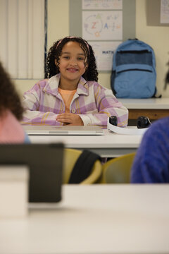 African American child sitting and smiling at school desk in pink jacket with closed laptop headset