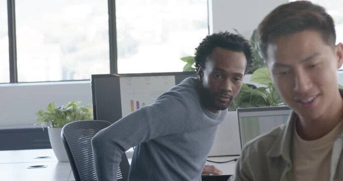 Diverse male team at desk, Asian coworker gesturing, African colleague rising, reviewing screen