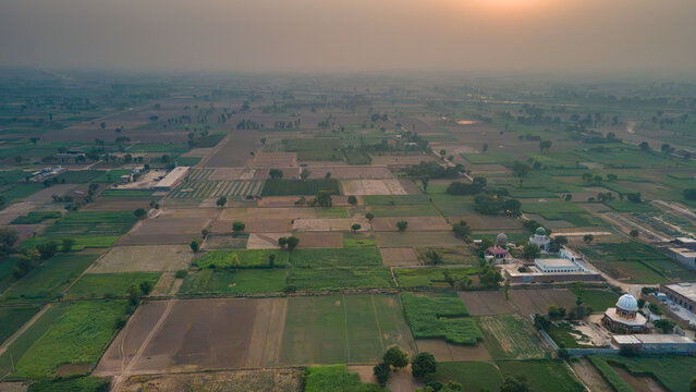 Aerial view of agricultural fields with green crops and patches of soil under a hazy sunset sky in Punjab, Pakistan.