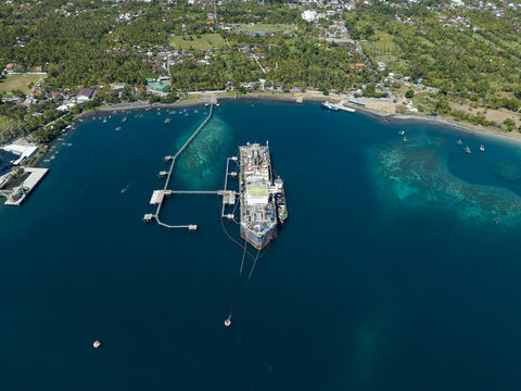 Aerial view of a Floating Storage and Offloading Ship moored in the clear blue waters of the bay, connected by a long jetty to the lush green coastline of Buleleng Regency, Bali, Indonesia.