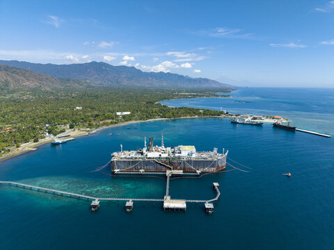 Aerial view of a Floating Storage and Offloading Ship anchored in clear blue waters with a long jetty connecting to the lush green shoreline of Buleleng Regency, Bali, Indonesia.