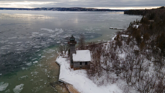Aerial view of Grand Island Lighthouse surrounded by snow-covered trees and icy waters of Lake Superior in Munising, Michigan, United States.