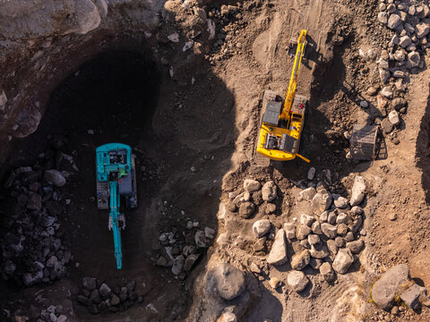 Aerial view of two excavators, one teal and one yellow, working in a rocky quarry with excavated earth and stone piles in Klaten, Central Java, Indonesia.