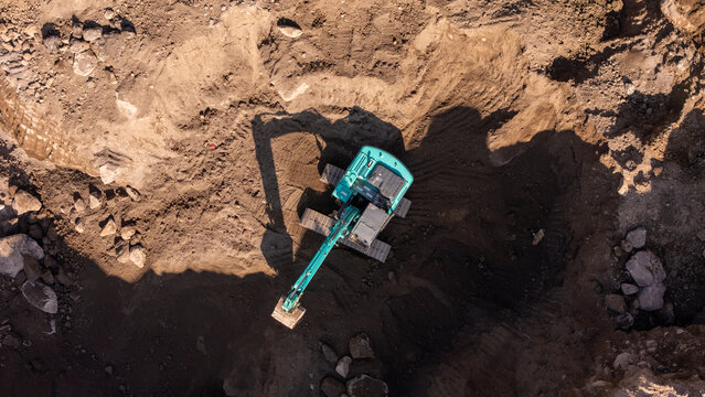 Aerial view of an excavator working in a deep dirt pit with rocky terrain and shadows cast on the ground in Klaten, Central Java, Indonesia.