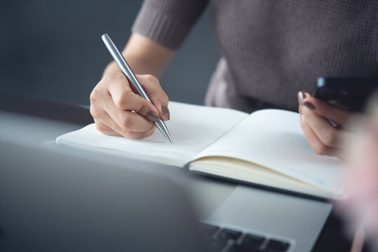 Woman using smartphone with laptop and writing on notebook, doing online research, remote working from home