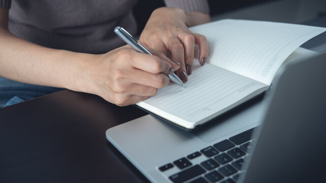 Woman hand with a pen writing on notebook with laptop on office desk, business planning. Student online studying and doing online research