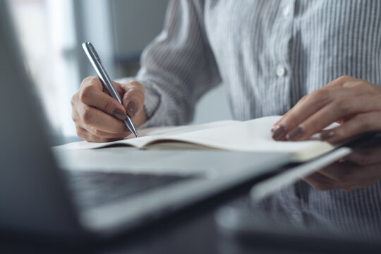 Close up of business woman hand with a pen writing on paper notebook with laptop on office desk. Business planning and reminder. Online research, searching on internet, e-learning