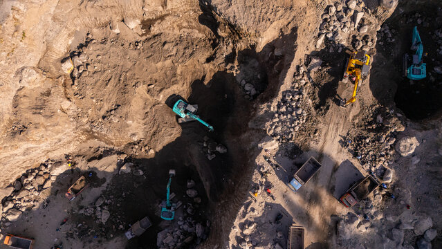 Aerial view of excavators and dump trucks working in a quarry with rocky terrain and piles of dirt in Klaten, Central Java, Indonesia.