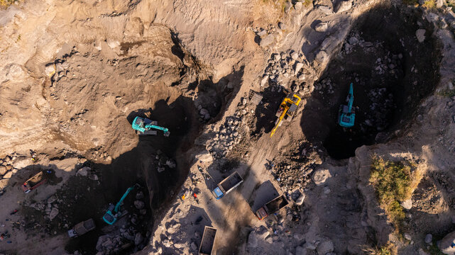 Aerial view of excavators and dump trucks working in a stone quarry with rocky terrain and deep pits in Klaten, Central Java, Indonesia.