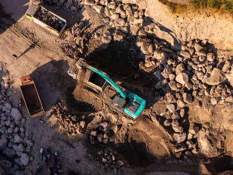 Aerial view of an excavator loading rocks into a dump truck at a mining quarry with workers nearby in Klaten, Central Java, Indonesia.