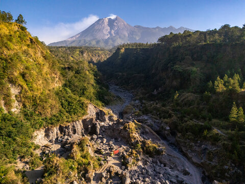Aerial view of Mount Merapi volcano rising above a rocky riverbed with lush green slopes and volcanic terrain in Klaten, Central Java, Indonesia.