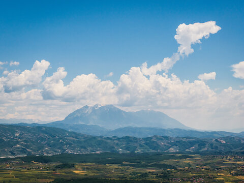 Aerial view of the mountainous landscape near Belesh, featuring rolling hills, fields, and a distant mountain range under a blue sky with clouds in Belesh, Qarku i Elbasanit, Albania.