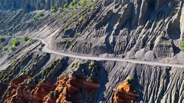 Aerial view of a steep dirt road carved into the side of an eroded canyon cliff with layered geological strata in Anjihai Grand Canyon, Xinjiang, China