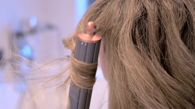 Woman using advanced hair styling tool to curl blonde locks for elegant hairstyle in bright bathroom.