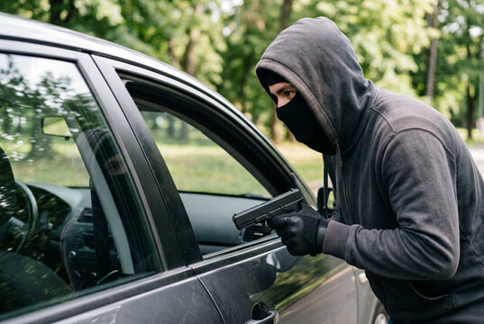 Masked criminal in gray hoodie aiming handgun at a car driver window